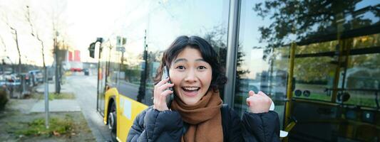Happy asian girl talks on mobile phone and looks surprised, amazed by great news, achieve goal, hear something exciting photo