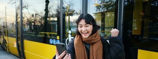 retrato de alegre asiático niña negociaciones en móvil teléfono, vídeo charlas, mira asombrado a teléfono inteligente cámara, soportes en autobús detener foto