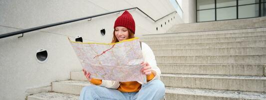 Beautiful girl tourist sits on stairs with city map, plans her journey, looks for direction while travelling around town, searches route for sightseeing photo