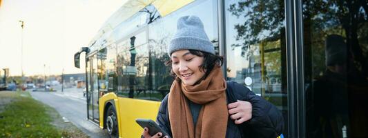 imagen de niña estudiante esperando para público transporte, cheques calendario en teléfono inteligente aplicación, soportes cerca ciudad autobús foto