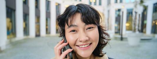 retrato de sonriente asiático niña negociaciones en móvil teléfono, habla con amigo en teléfono inteligente, camina en ciudad centro, soportes en calle y se ríe foto