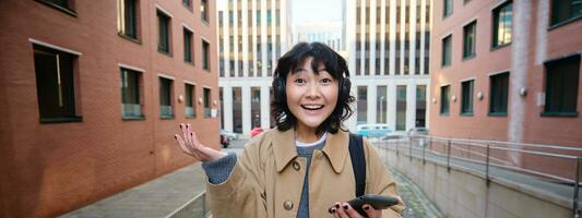 Portrait of young korean woman in headphones, looks amazed and surprised, hold mobile phone, realise something fantastic happened, cheering, celebrating victory photo