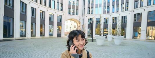 celular tecnología. joven coreano mujer negociaciones en móvil teléfono, hace un teléfono llamada en su camino hogar, camina abajo calle, ciudad centro, tiene teléfono conversacion foto