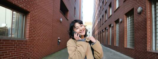 retrato de joven coreano mujer caminando abajo calle con teléfono, hablando con alguien, hace un llamar, tiene teléfono conversacion foto