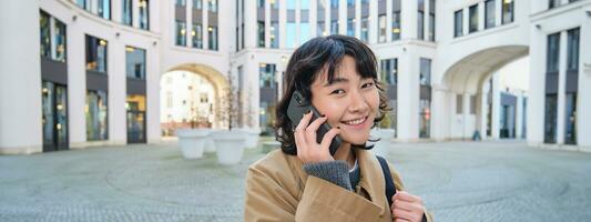 Cellular technology. Young korean woman talks on mobile phone, makes a phone call on her way home, walks down street, city centre, has telephone conversation photo