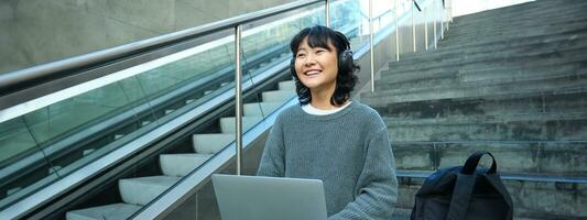 Female student works on project remotely, sits on stairs street, works on laptop, listens music in headphones and smiles photo