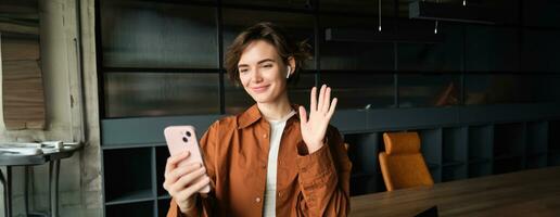 Image of woman connects to work call, video chat on mobile phone, waves hand at smartphone app and smiles, stands in an office in casual clothes photo