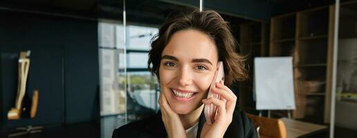Close up portrait of smiling saleswoman, office manager stands in conference room, answers phone call, talking on mobile telephone with someone, receives great news and rejoices photo