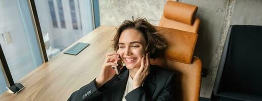 Portrait of corporate woman talking on mobile phone, answering a call in her office, laughing and smiling, having a conversation, leaning on her chair photo