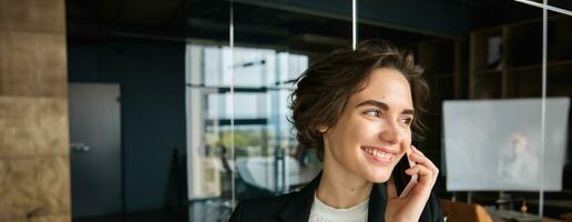 Corporate woman making phone calls, answering mobile and smiling, talking to client, having a conversation, standing in office in business suit photo