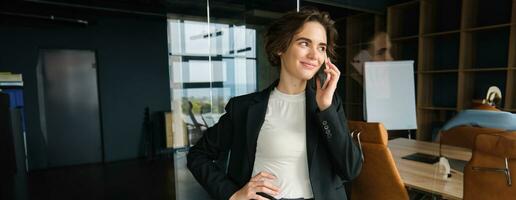 Image of confident businesswoman answering phone call, having a conversation over the telephone and smiling, standing in office or co-working space photo