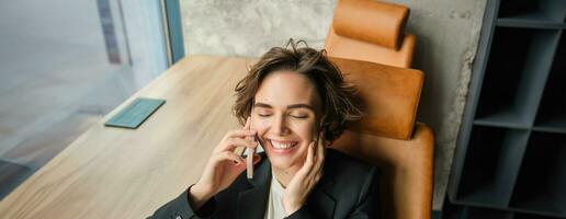 Portrait of business woman in suit, sitting in her office and answering a phone call with pleased smile, having a negotiation over the telephone conversation photo
