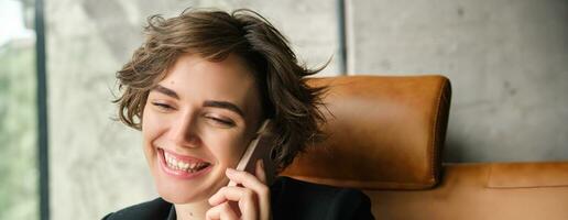 Corporate people and business concept. Young woman in suit, sitting in office and having conversation, answering phone call and smiling, talking to client photo