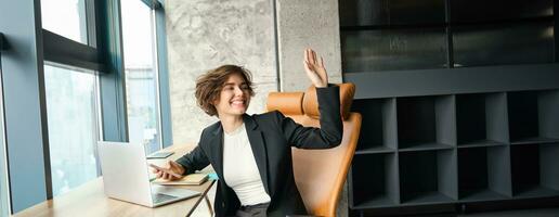 Portrait of young successful woman, corporate lady in an office, turn around and saying hello, waving hand to say hi to colleague, sitting with laptop and mobile phone photo