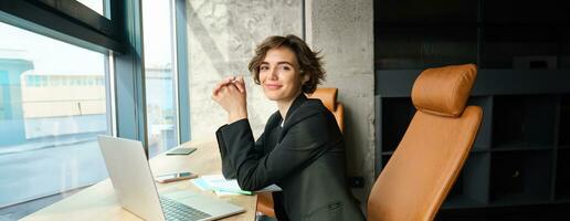 Image of corporate woman working in an office, sitting in front of laptop, preparing for business meeting, wearing black suit photo