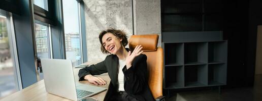 Cheerful businesswoman, office manager connects to video online meeting, saying hello and waving hand at laptop with friendly smiling face photo