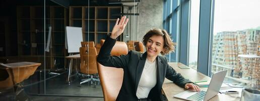 Portrait of woman working on laptop, businesswoman saying hello, waving at co-worker in her office and smiling, saying hello to colleague photo