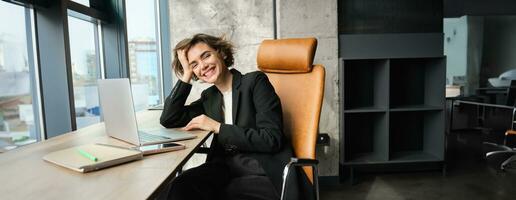 Portrait of young successful business woman in suit, sitting in office with laptop, smiling and looking happy, enjoys her job, working for company photo