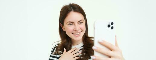 Portrait of happy woman taking photo, laughing while shooting something on smartphone, standing isolated over white background photo