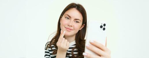 Portrait of beautiful woman showing heart sign and posing for selfie, taking picture of herself on smartphone app, posing near something cute, white background photo