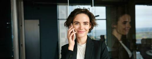 Portrait of successful businesswoman working in an office, standing near glass wall with mobile phone and smiling photo