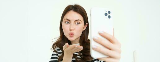 retrato de joven europeo mujer tomando selfie en teléfono inteligente, participación blanco móvil teléfono y posando para fotos, aislado en contra blanco antecedentes foto