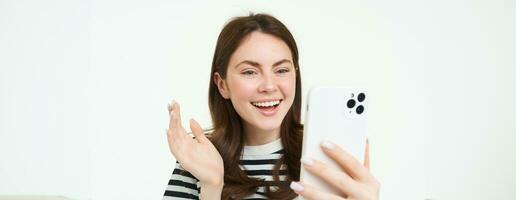 Portrait of young modern girl connects to video chats, talks with friend online using smartphone app, waves at mobile phone camera, white background photo