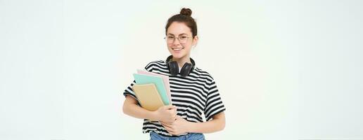 Education concept. Smiling brunette girl, student in casual clothes, holds her books, study material, wears headphones over neck, looks confident and happy, isolated over white background photo