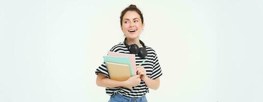 Student and education concept. Young woman with books, notes and pen standing over white background, college girl with headphones over neck posing in studio photo