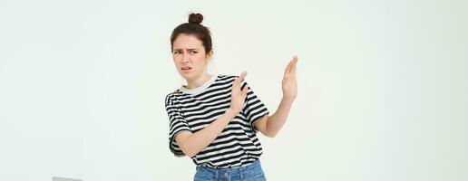 Image of woman expressing aversion, dislike something, looks apalled, extends hands forward in defensive gesture, rejecting, refusing, standing over white background photo