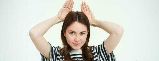 Image of cute brunette girl shows animal eats with hands on top of head, smiling and looking happy, white background photo