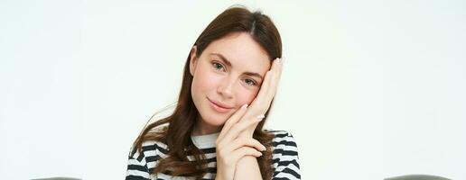 Portrait of tender, beautiful young woman, touches her clear, glowing skin, without blemishes or acne, concept of makeup and cosmetology, white background photo