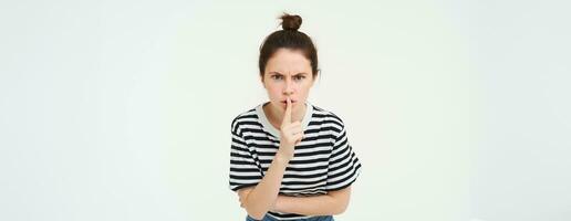 Image of angry woman shushing, makes hush gesture, press finger to lips with frustrated face, keep quiet, scolding loud person, white background photo