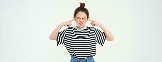Image of annoyed woman shuts her ears, cant stand noise, being disturbed by loud music, stands over white background photo