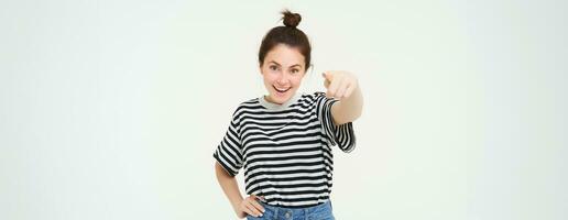 Image of confident, happy young woman in casual clothes, pointing finger at camera, laughing and smiling, standing over white background photo