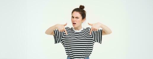 Frustrated girl points down, frowns and looks angry, asks questions with upset face expression, stands over white background photo