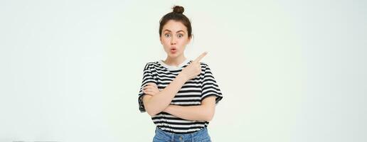 Image of girl with surprised face, looks amazed, points finger left, shows advertisement, white background photo