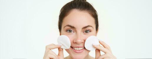 Close-up portrait of young woman washing her face, takes off her makeup with cotton pads, using facial toner, skincare cleanser, standing over white background photo