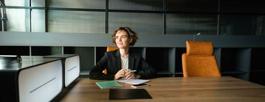 Young businesswoman waiting for start of meeting. Corporate woman in suit sitting in office, working on documents, waiting for client photo