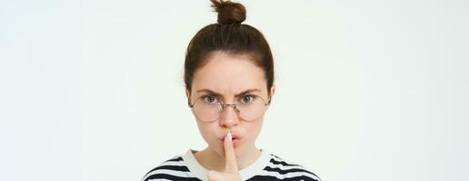 Image of serious woman in glasses, teacher shushing, makes shush, hush gesture, press finger to lips and frowning from disapproval, standing over white background photo