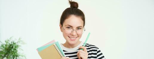 Close up portrait of young woman, tutor, online teacher in glasses, holding pen and documents, carries notebooks, stands over white background photo