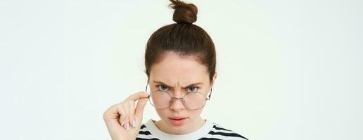 Portrait of angry woman looks from under forehead, takes off her glasses and stares annoyed at camera, stands over white background photo