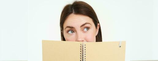 Young woman holds notebook daily planner next to her face, writing down homework, making notes, looking thoughtful, standing over white background photo