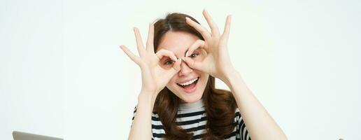 Portrait of happy, beautiful young woman, makes glasses with fingers around eyes and laughing, having fun, enjoying event, standing over white background photo