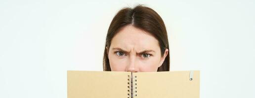 Woman with confused face looking at camera, frowning, holding notebook, reading planner or personal diary, standing over white background photo