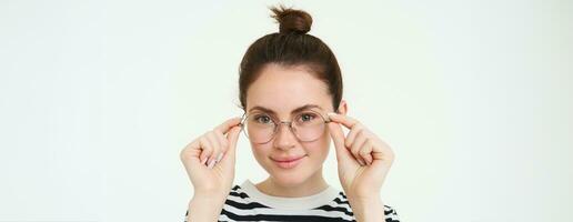 Close up portrait of smiling, attractive young woman wearing new eyewear, trying on glasses for vision, standing over white background photo