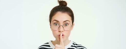 Portrait of girl with glasses, looks surprised, hughing, press finger to lips, shush shh gesture, asks to keep quiet, stands over white background photo