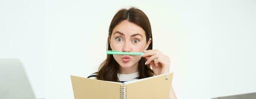 Portrait of funny, smiling woman with notebook, holding pen next to her lip and looking happy, white background photo