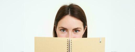 Portrait of cute young woman hides her face behind planner, holds notebook against her face and smiles, isolated over white background photo