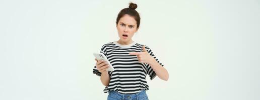 Image of angry young woman complains, points at telephone and looks disappointed, isolated against white background photo